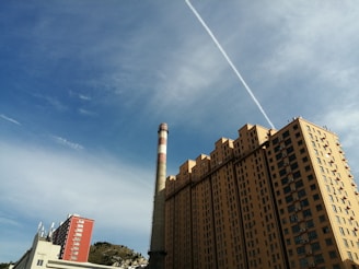 A tall residential building with multiple balconies and a large chimney nearby. The sky is mostly clear with a prominent contrail running diagonally across. In the distance, there is a small hill with sparse vegetation. Another smaller red building is also visible.