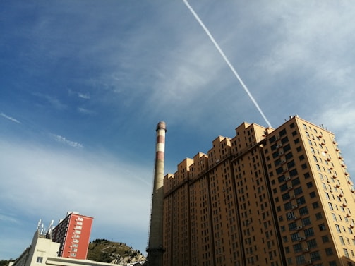A tall residential building with multiple balconies and a large chimney nearby. The sky is mostly clear with a prominent contrail running diagonally across. In the distance, there is a small hill with sparse vegetation. Another smaller red building is also visible.