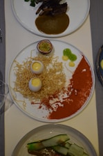 A gourmet dish consisting of a white plate featuring a half passion fruit, crispy noodles, two egg-shaped items with a creamy yellow center, dots of green gel, a red powder spread across the plate, and another dish with ribs and sauce in the background. There is also a dish with zucchini strips in a sauce.