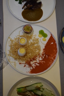A gourmet dish consisting of a white plate featuring a half passion fruit, crispy noodles, two egg-shaped items with a creamy yellow center, dots of green gel, a red powder spread across the plate, and another dish with ribs and sauce in the background. There is also a dish with zucchini strips in a sauce.