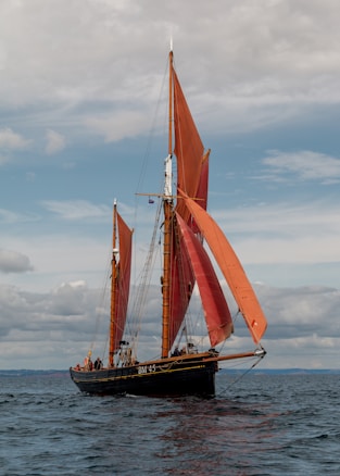 A classic sailing vessel with tall, reddish-brown sails glides on a calm sea under a partly cloudy sky. The sails are fully unfurled, catching the wind as the ship moves forward. Several people are visible on the deck, engaging with the ship's rigging and other activities. The background consists of the open sea stretching to the horizon and a sky filled with a mix of clouds.