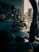 A motorcycle showroom with a vintage motorcycle displayed in the foreground. Various motorcycle parts, including wheels and a seat, are mounted on the wall as decor. The area is well-lit with a modern urban view visible through the window.