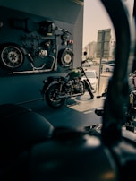 A motorcycle showroom with a vintage motorcycle displayed in the foreground. Various motorcycle parts, including wheels and a seat, are mounted on the wall as decor. The area is well-lit with a modern urban view visible through the window.