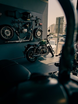 A motorcycle showroom with a vintage motorcycle displayed in the foreground. Various motorcycle parts, including wheels and a seat, are mounted on the wall as decor. The area is well-lit with a modern urban view visible through the window.