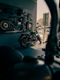 A motorcycle showroom with a vintage motorcycle displayed in the foreground. Various motorcycle parts, including wheels and a seat, are mounted on the wall as decor. The area is well-lit with a modern urban view visible through the window.