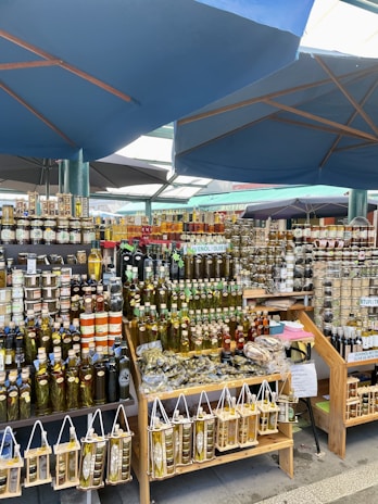 A market stall features a variety of bottles filled with olive oil and other condiments, neatly arranged on wooden shelves. Numerous jars of preserves and other products are stacked in the background. The setup is beneath large blue umbrellas in an outdoor market setting.