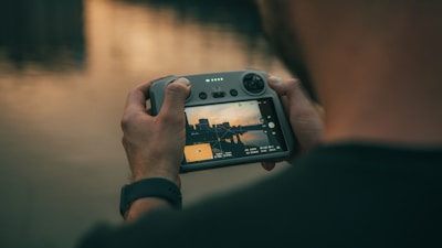 A person is holding a remote control device with a screen displaying a live video feed of a cityscape at sunset. The controls and buttons of the device are clearly visible. The background shows a blurred water surface reflecting the colors of the sunset.