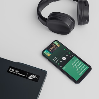 A set of black over-ear headphones, a smartphone displaying a music streaming app with a playlist or song in progress, and a closed laptop are arranged on a light-colored surface. The smartphone shows text in a non-English script and album art for a song.