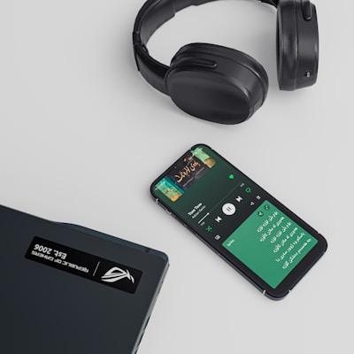 A set of black over-ear headphones, a smartphone displaying a music streaming app with a playlist or song in progress, and a closed laptop are arranged on a light-colored surface. The smartphone shows text in a non-English script and album art for a song.