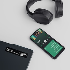 A set of black over-ear headphones, a smartphone displaying a music streaming app with a playlist or song in progress, and a closed laptop are arranged on a light-colored surface. The smartphone shows text in a non-English script and album art for a song.