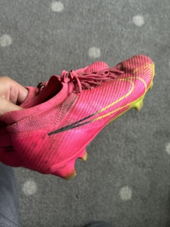 A hand is holding a bright pink soccer cleat with black and yellow detailing. The shoe features a textured surface, lace-up design, and studded sole, suitable for playing on grass. The background shows a gray carpet with lighter circular patterns.