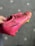 A hand is holding a bright pink soccer cleat with black and yellow detailing. The shoe features a textured surface, lace-up design, and studded sole, suitable for playing on grass. The background shows a gray carpet with lighter circular patterns.