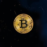 A golden coin featuring the Bitcoin symbol is centered against a black background. The coin displays intricate circuit-like patterns and inscriptions around its edge.
