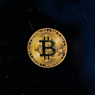 A golden coin featuring the Bitcoin symbol is centered against a black background. The coin displays intricate circuit-like patterns and inscriptions around its edge.