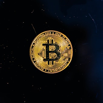 A golden coin featuring the Bitcoin symbol is centered against a black background. The coin displays intricate circuit-like patterns and inscriptions around its edge.