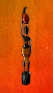 A wooden sculpture of a stylized human figure with elongated features and minimalist design. The figure stands on a black wooden base, with one arm raised, holding a red object that resembles a flame or leaf. The sculpture exhibits smooth textures and subtle color variations in wood tones.