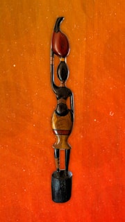 A wooden sculpture of a stylized human figure with elongated features and minimalist design. The figure stands on a black wooden base, with one arm raised, holding a red object that resembles a flame or leaf. The sculpture exhibits smooth textures and subtle color variations in wood tones.