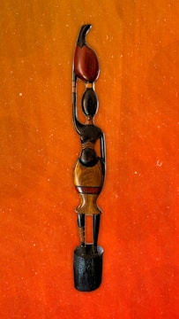 A wooden sculpture of a stylized human figure with elongated features and minimalist design. The figure stands on a black wooden base, with one arm raised, holding a red object that resembles a flame or leaf. The sculpture exhibits smooth textures and subtle color variations in wood tones.