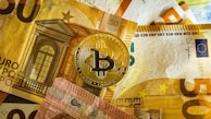 A golden Bitcoin physical coin is placed on top of several Euro banknotes. The banknotes display architectural imagery and a mix of colors including yellow, brown, and red, with the Bitcoin logo prominently visible on the coin.