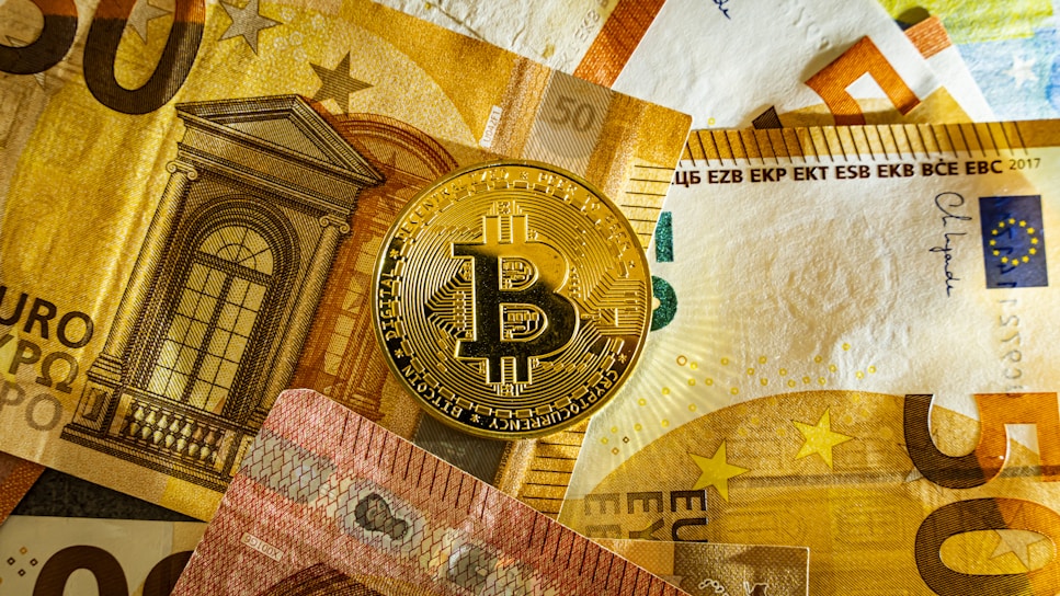 A golden Bitcoin physical coin is placed on top of several Euro banknotes. The banknotes display architectural imagery and a mix of colors including yellow, brown, and red, with the Bitcoin logo prominently visible on the coin.