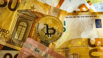 A golden Bitcoin physical coin is placed on top of several Euro banknotes. The banknotes display architectural imagery and a mix of colors including yellow, brown, and red, with the Bitcoin logo prominently visible on the coin.