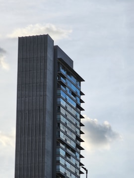 A modern high-rise building with a distinctive architectural design stands tall against a backdrop of a partly cloudy sky. The structure features a concrete facade on one side and glass windows that reflect the sky on the other. The lines are clean and emphasize a contemporary aesthetic.