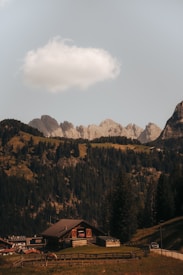 A serene mountain landscape with a wooden cabin situated in the foreground. The scene features lush green pine forests covering the mountainous terrain. A single white cloud floats above the jagged mountain peaks in the clear blue sky. There is a fenced area near the cabin with a grazing horse.
