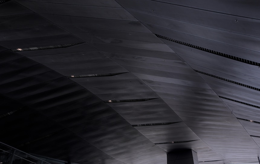 A detailed view of a modern architectural ceiling made up of overlapping dark metal panels with linear vents. The panels create geometric patterns with subtle variations in shading due to lighting.