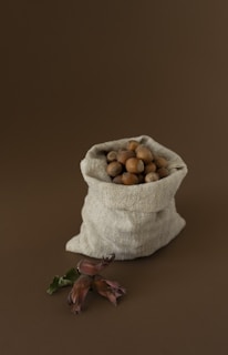 A rustic burlap sack filled with hazelnuts is placed against a brown background. In front of the sack, a branch with hazelnuts and leaves is displayed, adding a natural element to the composition.
