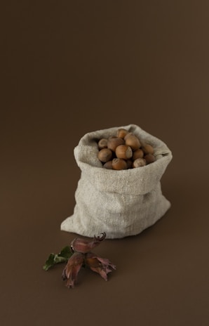 A rustic burlap sack filled with hazelnuts is placed against a brown background. In front of the sack, a branch with hazelnuts and leaves is displayed, adding a natural element to the composition.