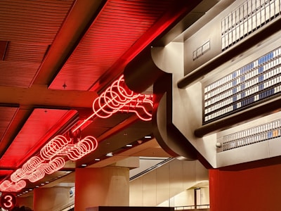 A red neon light installation is mounted on the ceiling of a modern interior space with industrial design elements. The lights form a geometric pattern with circular shapes, casting a warm glow. On one side, metallic architectural features are visible, resembling a grid-like structure, providing a contrast to the warm lighting.