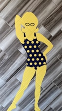 A silhouette of a person wearing a black polka dot swimsuit with large yellow polka dots, standing on a tiled floor. The silhouette is yellow and features accessories like sunglasses and hoop earrings. The background consists of gray wooden tiles arranged in a herringbone pattern.