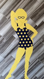 A silhouette of a person wearing a black polka dot swimsuit with large yellow polka dots, standing on a tiled floor. The silhouette is yellow and features accessories like sunglasses and hoop earrings. The background consists of gray wooden tiles arranged in a herringbone pattern.