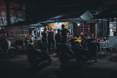 A bustling nighttime street food stall with several people gathered around. The stall is illuminated, showcasing signs with food offerings. Motorbikes are parked in front, and a few customers are seated at a table nearby.