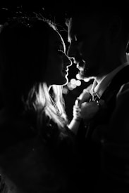 Two people are in silhouette, closely facing each other, possibly about to kiss. The scene is dark with a moody atmosphere and soft bokeh lights in the background.