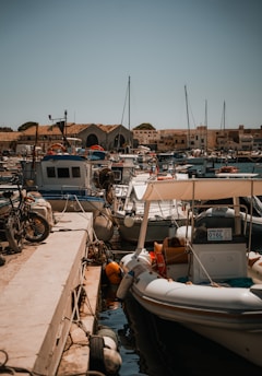 A bustling marina with numerous boats moored along a concrete dock. The scene includes sailboats and motorboats, with ropes and buoys scattered around. Bicycles are parked nearby, and buildings are visible in the background under a clear blue sky.