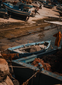 A collection of small, rustic fishing boats rests on a sandy and rocky shore. The vessels are primarily blue with weathered wood, suggesting they have been used extensively. Fishing nets and ropes are visible, hinting at their function. The sunlight casts a warm glow across the scene, adding a nostalgic, maritime feel.