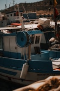 A small fishing boat docked at a harbor, marked with the name 'MIRIAM LTP403.' The boat features blue and white paint and is equipped with a fishing reel. Surrounding the boat, there are other vessels, some with flags. In the background, a rocky hillside is visible under a clear sky.