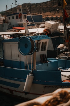 A small fishing boat docked at a harbor, marked with the name 'MIRIAM LTP403.' The boat features blue and white paint and is equipped with a fishing reel. Surrounding the boat, there are other vessels, some with flags. In the background, a rocky hillside is visible under a clear sky.