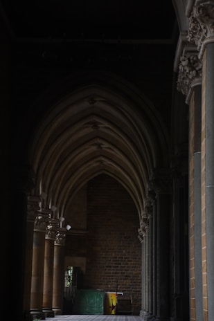 Gothic-style arches with intricate stone carvings line a corridor with a brick wall at the far end. The setting is dimly lit, creating deep shadows and a mysterious ambiance. Several tall, narrow columns support the arches, and there appears to be some stored items in the corner near a window.