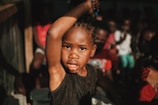 A child in focus with raised arm and an intense expression, surrounded by other children in the background. The lighting creates contrast and emphasizes the child's features.