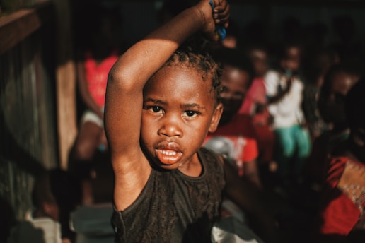 A child in focus with raised arm and an intense expression, surrounded by other children in the background. The lighting creates contrast and emphasizes the child's features.