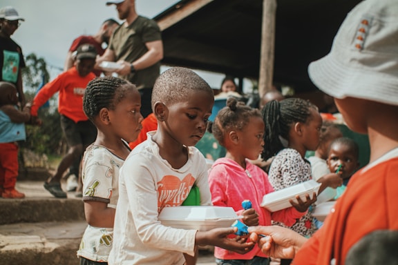 A group of children and a few adults are gathered outdoors, with some children holding food containers. They appear to be waiting in line or engaging in a communal activity. The setting looks informal, possibly in a rural or community space.