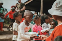 A group of children and a few adults are gathered outdoors, with some children holding food containers. They appear to be waiting in line or engaging in a communal activity. The setting looks informal, possibly in a rural or community space.
