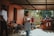 A group of people, including several children, are gathered outdoors under a covered area. A woman stands in the foreground, speaking or presenting to the group. The setting appears to be a residential area with a concrete floor and orange walls. The background includes greenery, indicating an outdoor environment.