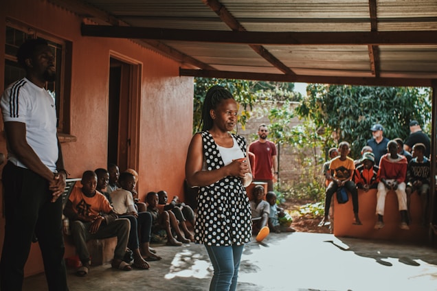 A group of people, including several children, are gathered outdoors under a covered area. A woman stands in the foreground, speaking or presenting to the group. The setting appears to be a residential area with a concrete floor and orange walls. The background includes greenery, indicating an outdoor environment.
