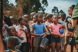 A group of children standing outside, some holding small bags and a red notebook. They are gathered closely together with some wearing casual clothing like t-shirts and shorts. The background shows trees and a clear sky.