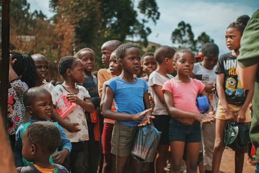 A group of children standing outside, some holding small bags and a red notebook. They are gathered closely together with some wearing casual clothing like t-shirts and shorts. The background shows trees and a clear sky.