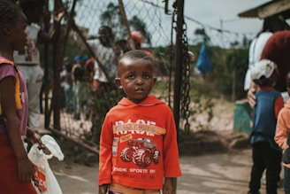 A young child wearing a red sweater stands in the foreground with a neutral expression, surrounded by other children and a chain-link fence in the background. The scene appears to be outdoors, capturing a moment in what seems to be a community or school setting.