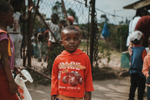 A young child wearing a red sweater stands in the foreground with a neutral expression, surrounded by other children and a chain-link fence in the background. The scene appears to be outdoors, capturing a moment in what seems to be a community or school setting.